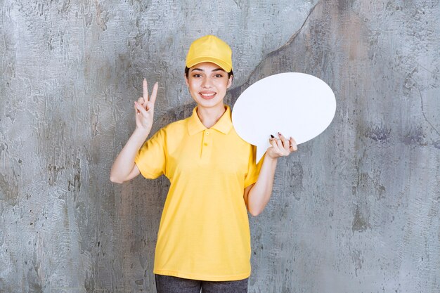 Agent de service féminin en uniforme jaune tenant un panneau d'information ovale et montrant un signe positif de la main.