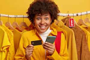 Photo gratuite agréable femme afro vérifie le compte bancaire, paie en ligne via smartphone, détient une carte de crédit