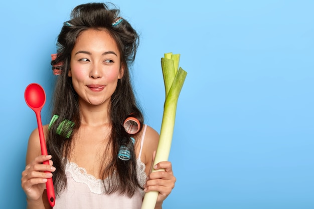 Agréable femme au foyer brune avec apparence asiatique, tient une cuillère et un poireau vert, prépare un petit-déjeuner végétarien