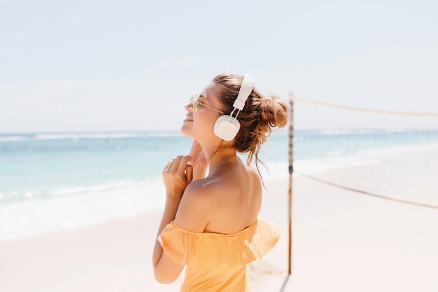 Agréable femme caucasienne s'amusant sur la côte de l'océan et écoutant de la musique Jolie fille en tenue orange passant le matin d'été en mer