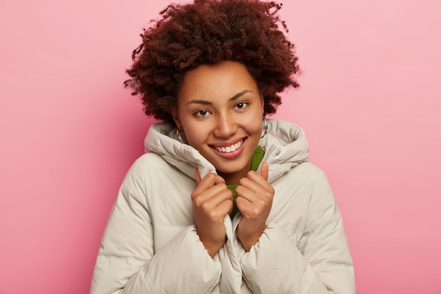 Agréable femme heureuse avec des cheveux bouclés, porte un manteau chaud et confortable, a une peau foncée saine, sourit agréablement, isolée sur fond rose.