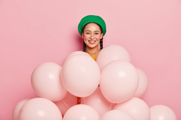 Agréable Modèle Féminin Asiatique Porte Un Béret Vert, Se Tient Près De Nombreux Ballons, Pose Sur Fond Rose, Fête Son Anniversaire