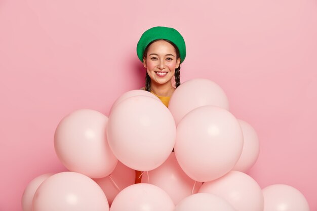 Agréable modèle féminin asiatique porte un béret vert, se tient près de nombreux ballons, pose sur fond rose, fête son anniversaire