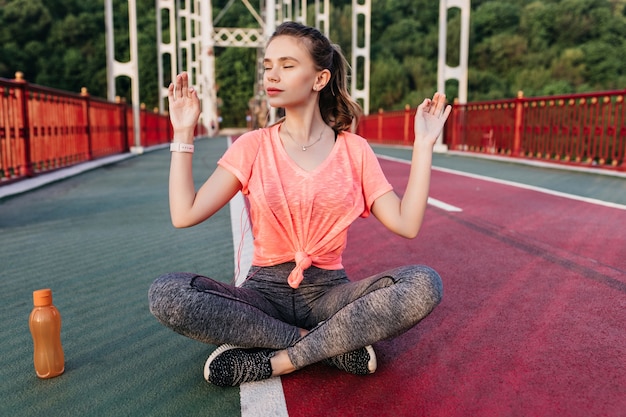 Agréable modèle féminin blanc se détendre après un entraînement intensif. Tir extérieur d'une charmante femme en baskets noires faisant du yoga au stade.