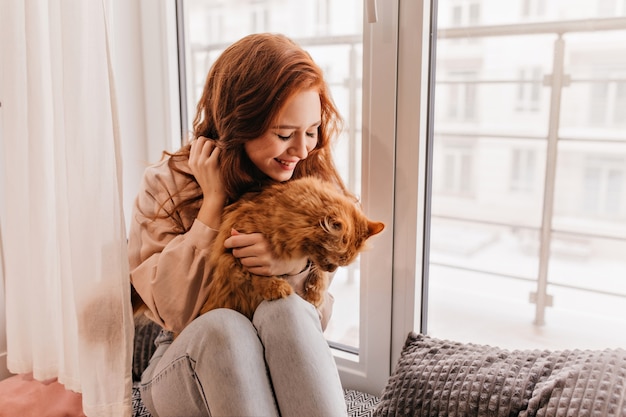 Agréable modèle féminin tenant un chat rouge sur ses genoux. Portrait intérieur de charmante femme au gingembre posant avec animal de compagnie.