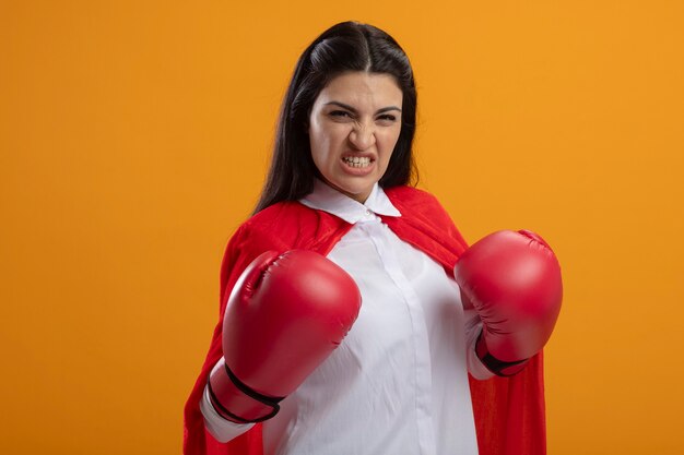 Agressif jeune superwoman portant des gants de boîte à l'avant en gardant les mains dans l'air isolé sur mur orange