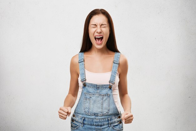 Agressivité, réaction humaine négative et attitude. Plan intérieur d'une jeune femme stressée en colère criant à haute voix avec la bouche grande ouverte, les yeux fermés et les poings serrés, ayant un regard fou et furieux