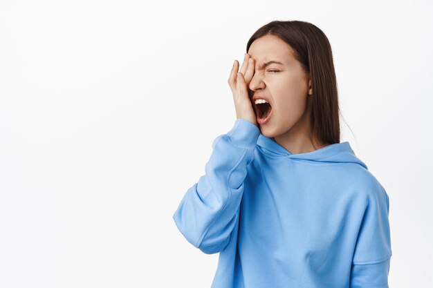 Aïe, ça fait mal. Jeune femme se frotte les yeux et se plaint, sentiment d'inconfort douloureux, debout dans un sweat à capuche bleu sur fond blanc. Copier l'espace