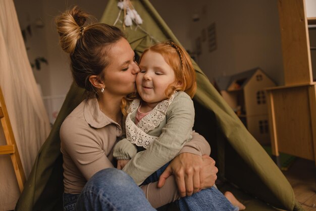 Aimer la jeune femme à la peau claire mère embrasse son enfant tenant une fille dans les bras tout en étant assis dans la chambre