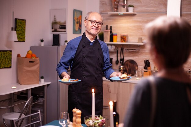 Aîné âgé servant le dîner à sa femme tout en ayant un dîner romantique. Couple de personnes âgées parlant, assis à table dans la cuisine, savourant le repas, célébrant leur anniversaire dans la salle à manger.