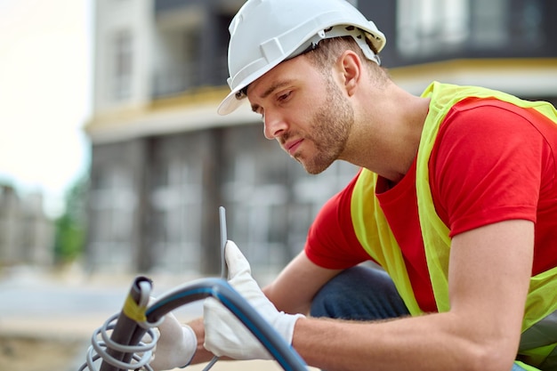 Ajustement. Jeune homme adulte portant des gants de casque de protection et un gilet lumineux accroupi regardant l'outil sur le côté de la caméra sur le chantier de construction