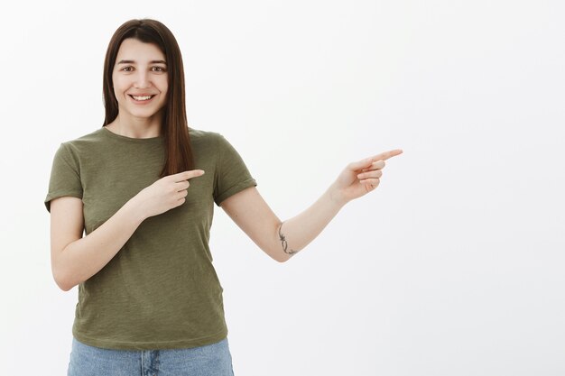 Allez je te montre. Enthousiaste jeune femme ordinaire lumineuse et heureuse avec tatouage et cheveux bruns souriant ravi et sympathique comme pointant vers la droite, indiquant à un endroit frais, voie de premier plan