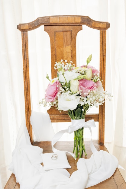 Photo gratuite alliances sur un coussin blanc avec écharpe et bouquet sur la chaise en bois près du rideau