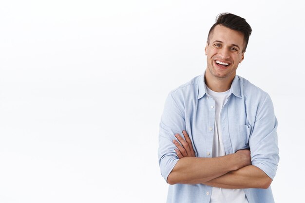 Allons droit au but. Portrait d'un jeune homme riant insouciant et détendu, bras croisés sur la poitrine dans une pose professionnelle prête, enthousiaste, gérant son propre petit magasin, mur blanc