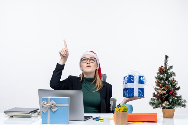 Ambiance festive avec curieuse jeune femme positive avec chapeau de père Noël et portant des lunettes assis à une table montrant un cadeau de Noël pointant au-dessus sur fond blanc