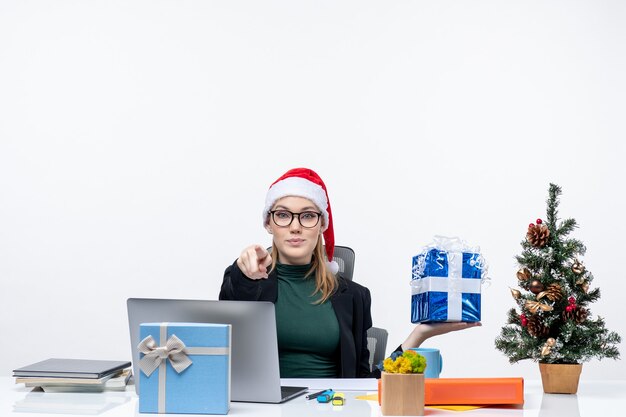 Ambiance festive de Noël avec curieux jeune femme positive avec chapeau de père Noël et portant des lunettes assis à une table montrant un cadeau pointant quelqu'un sur fond blanc