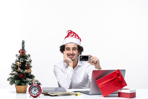 Ambiance festive de vacances avec homme d'affaires souriant avec chapeau de père Noël tenant une carte bancaire et mettant sa main sous le menton au bureau