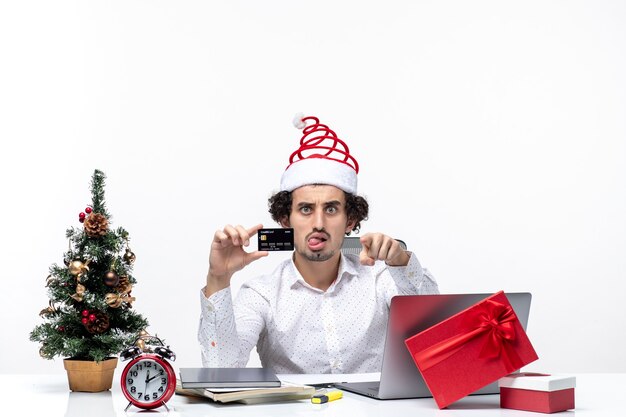 Ambiance festive de vacances avec jeune homme d'affaires avec chapeau de père Noël et tenant sa carte bancaire et pointant quelque chose dans le bureau sur fond blanc