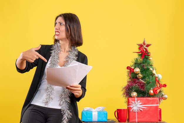 Ambiance de Noël avec belle dame confuse debout dans le bureau et pointant des documents dans le bureau sur jaune