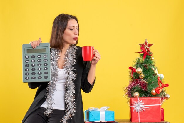 Ambiance de Noël avec une belle dame souriante debout dans le bureau et tenant une tasse de calculatrice au bureau sur jaune