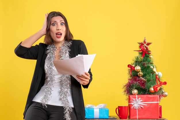 Ambiance de Noël avec une belle dame surprise émotionnelle debout dans le bureau et tenant des documents au bureau sur jaune