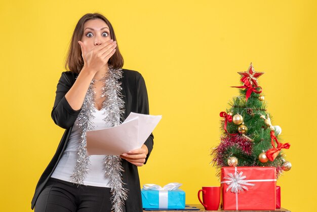 Ambiance de Noël avec surprise belle dame debout dans le bureau et tenant des documents au bureau sur jaune