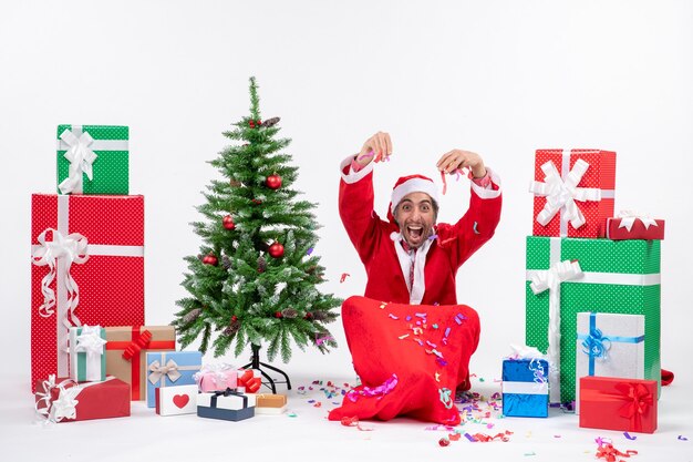 Ambiance de vacances festives avec drôle de père Noël heureux positif assis sur le sol et jouant avec des décorations de Noël près de cadeaux et arbre de Noël décoré sur fond blanc