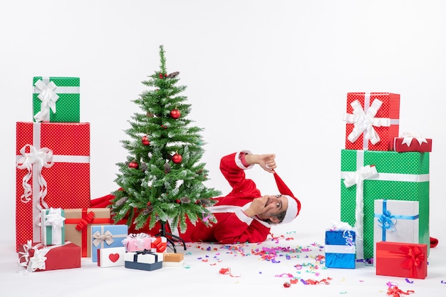 Ambiance de vacances festives avec jeune père Noël surpris couché derrière l'arbre de Noël près de cadeaux sur fond blanc