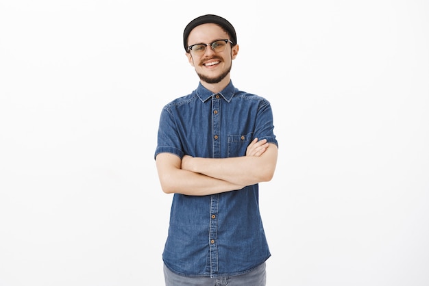 Ambitieux, créatif et beau mec mature avec une moustache en bonnet noir et des lunettes tenant les mains croisées sur la poitrine avec un sourire confiant et sûr de lui posant