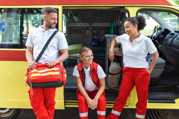 Ambulanciers paramédicaux et médecin debout sur le côté ambulance Le médecin porte un sac de traumatologie médicale Groupe de trois ambulanciers paramédicaux debout devant l'ambulance avec le sourire