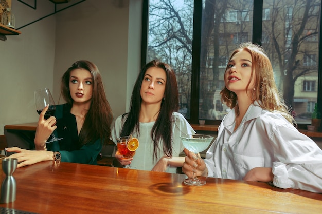 Amies ayant un verre au bar. Ils sont assis à une table en bois avec des cocktails. Ils portent des vêtements décontractés.