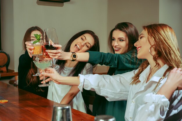 Amies ayant un verre au bar. Ils sont assis à une table en bois avec des cocktails. Ce sont des verres cliquetants