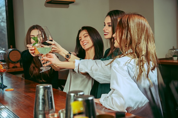 Amies ayant un verre au bar. Ils sont assis à une table en bois avec des cocktails. Ce sont des verres qui tintent