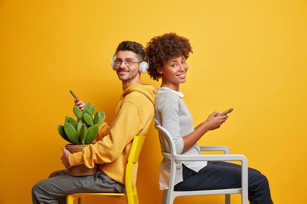 Des amis interraciaux posent sur des chaises contre un mur jaune vif, tiennent les téléphones portables et regardent avec des expressions joyeuses