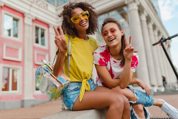 Amis de jeunes filles heureux élégantes colorées souriant assis dans la rue, les femmes s'amusant ensemble