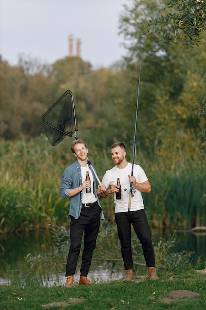 Photo gratuite des amis se tiennent près de la rivière et pêchent. deux pêcheurs discutant et buvant une bière