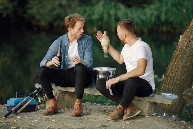 Photo gratuite des amis sont assis près d'un arbre et pêchent. deux pêcheurs discutant et buvant une bière