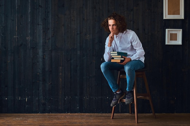 Photo gratuite l'amour de l'éducation. un jeune étudiant pensif tient des livres assis sur une chaise dans une pièce à l'intérieur minimaliste.