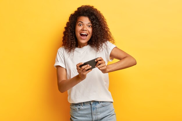 Amusé jeune femme aux cheveux bouclés posant avec son téléphone