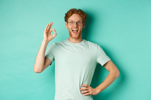 Amusé jeune homme aux cheveux rouges, portant des lunettes et un t-shirt, montrant un signe correct et souriant excité, vérifiant quelque chose et l'approuvant, debout sur fond turquoise.