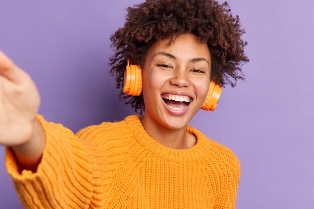 Amusée belle femme afro-américaine s'étire le bras prend selfie rit joyeusement aime écouter de la musique via des écouteurs sans fil porte des poses de chandail tricoté orange