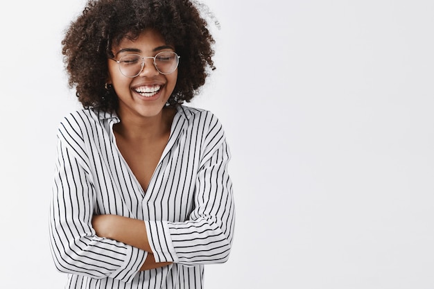 Amusée et insouciante jolie femme afro-américaine en chemisier rayé et lunettes fermant les yeux rire aux éclats et se tenant la main sur la poitrine fermant les yeux s'amusant