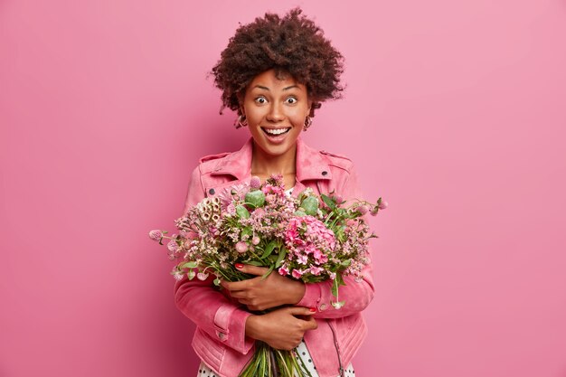 Amusée surprise, la femme à la peau sombre embrasse le bouquet de jolies fleurs, va féliciter un ami avec son anniversaire, porte une veste à la mode rose, se tient à l'intérieur. Célébration, occasion spéciale