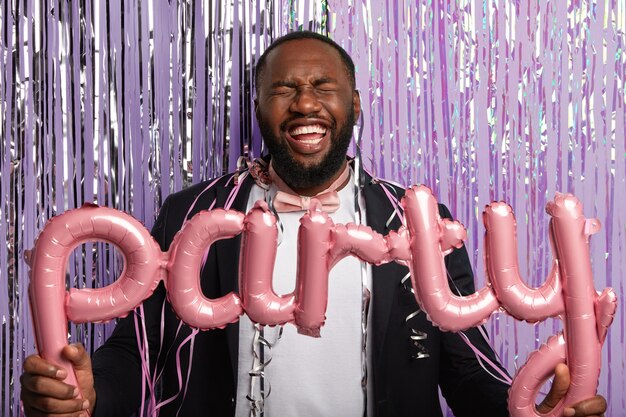 Amusement, bonheur, concept de vacances. Heureux marié à la peau sombre organise une fête de cerf, tient des ballons dans les mains, rit des émotions positives