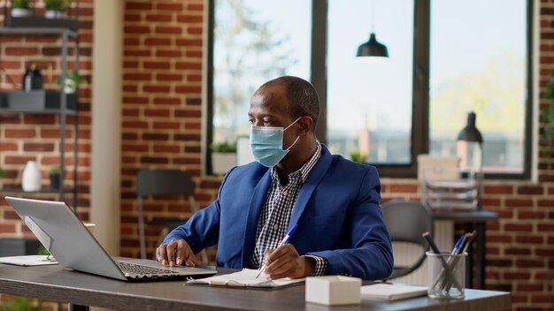 Analyste financier prenant des notes sur des documents commerciaux naviguant sur Internet sur un ordinateur portable, faisant de la paperasse pour planifier une stratégie. Homme d'affaires avec masque facial écrivant un rapport de statistiques. Prise de vue au trépied.