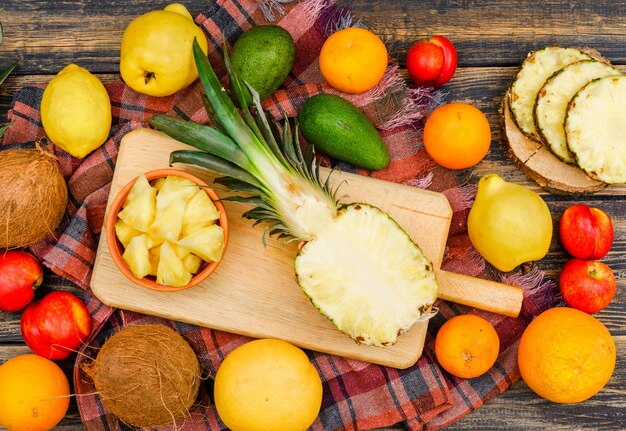 Ananas juteux en tranches dans des planches de bois et un bol avec des noix de coco, des pêches, des coings et des agrumes sur une surface grunge en bois et un tissu de pique-nique