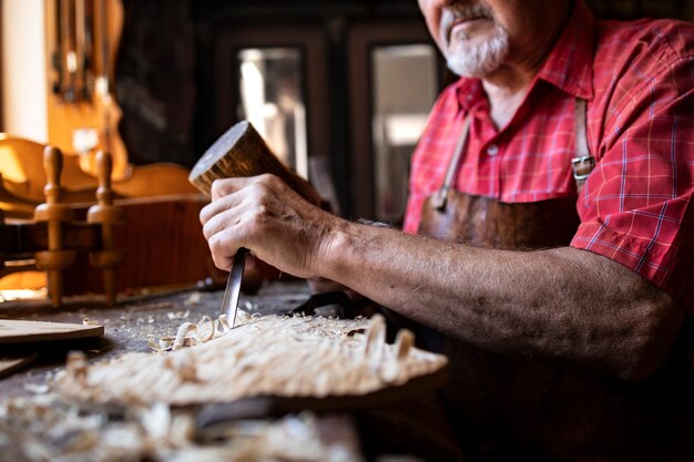 Un ancien menuisier senior expérimenté tenant un couteau et un marteau de sculpture sur planche de bois dans son atelier de menuiserie