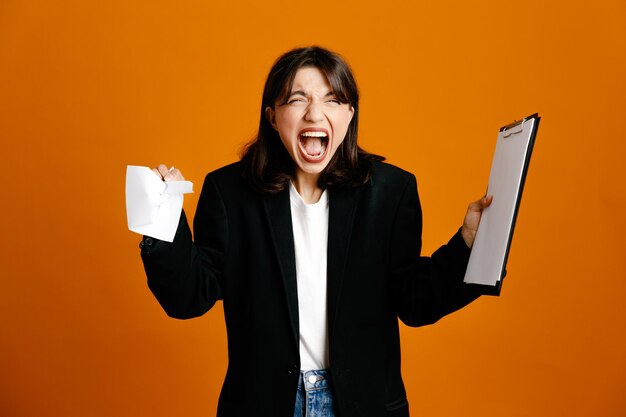 Angry holding presse-papiers avec du papier belle jeune femme portant une veste noire isolée sur fond orange