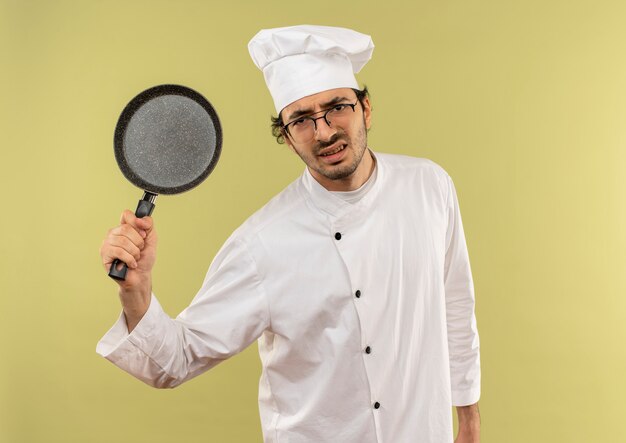 Angry young male cook portant l'uniforme de chef et des verres soulevant une poêle à frire