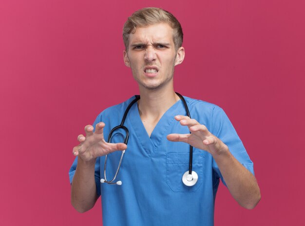 Angry young male doctor wearing doctor uniform avec stéthoscope montrant le geste de style tigre isolé sur mur rose